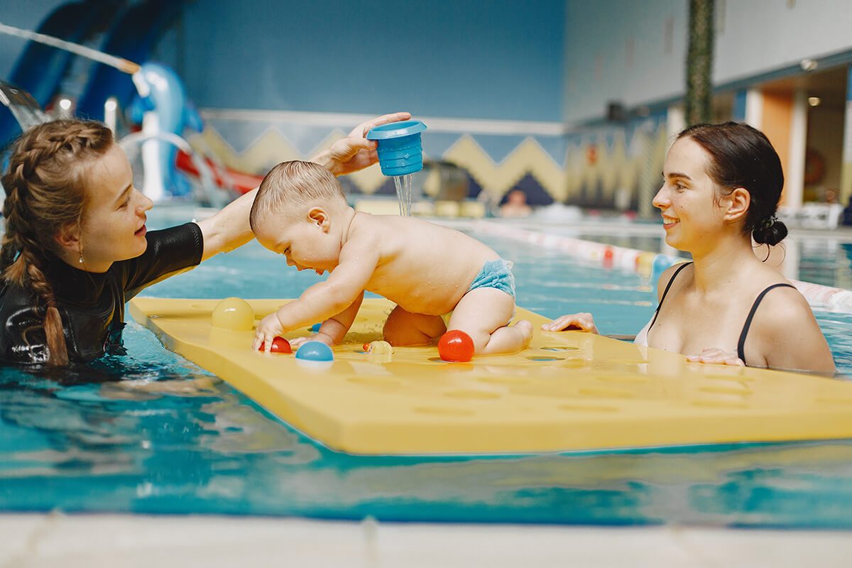 Un bébé qui s'amuse sur un tapis flottant avec sa maman et une maitre-nageur