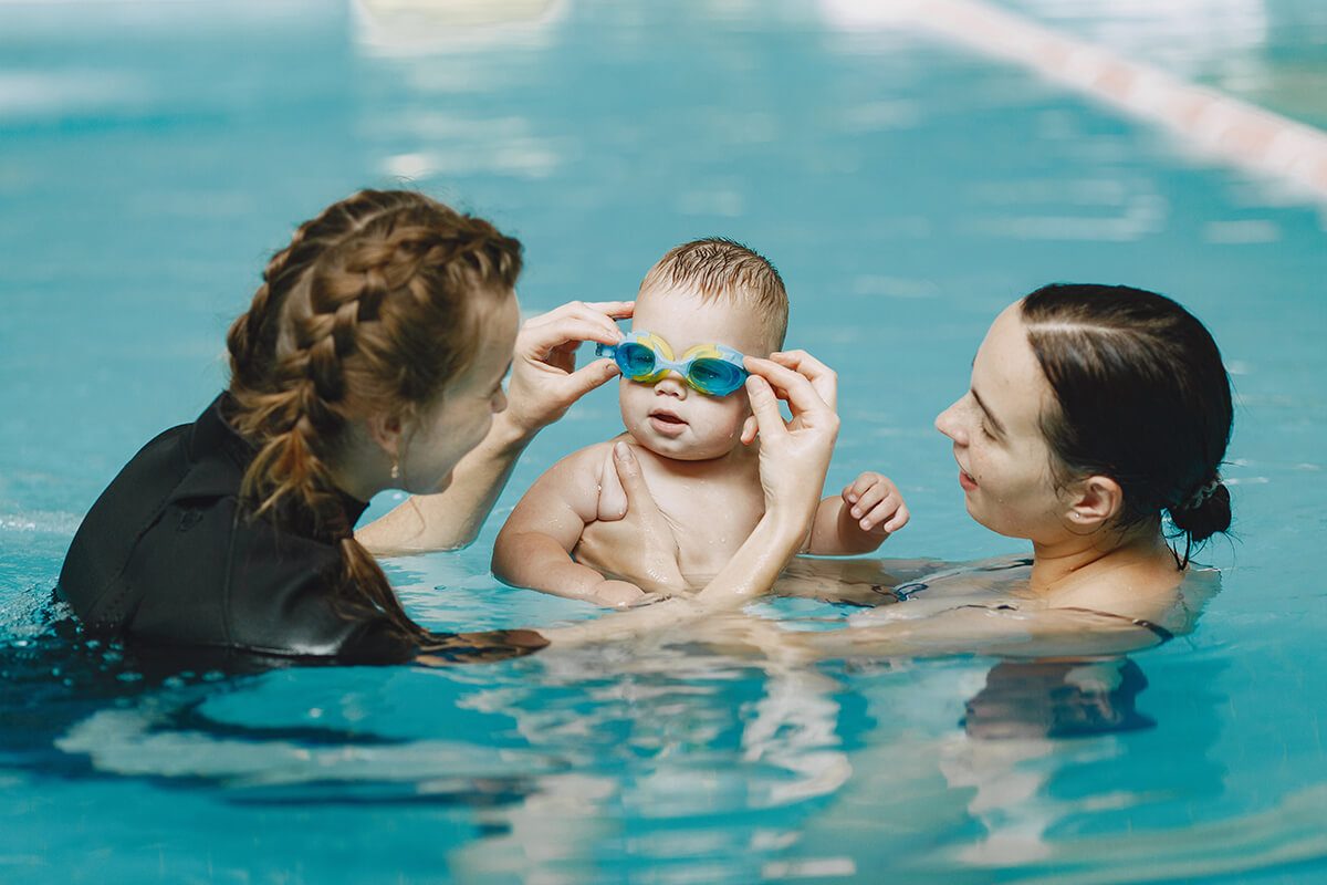 Un bébé avec des lunettes de piscine accompagné de sa maman et d'une maitre-nageur