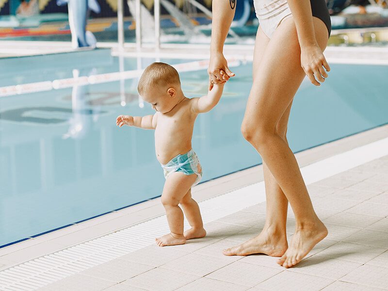 Un bébé avec une couche de piscine tenu par la main de sa maman au bord d'un bassin