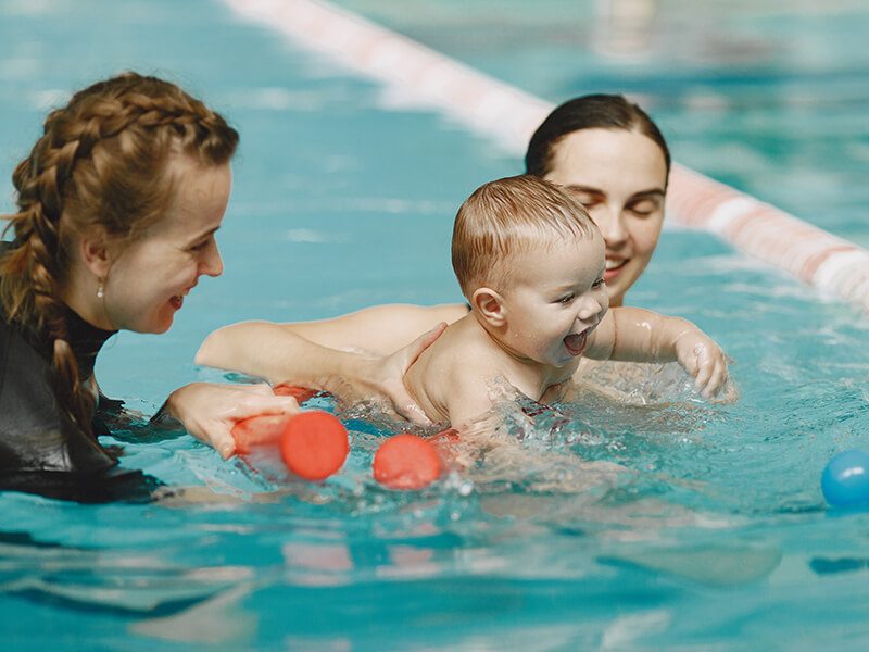 Un bébé qui joue dans l'eau entouré de sa maman et d'une maitre-nageuse à la piscine