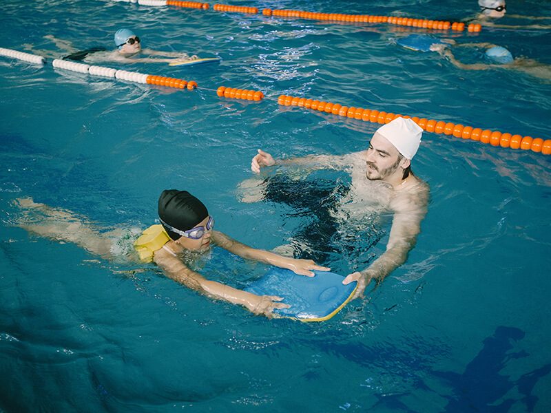 Un enfant qui apprend à nager avec une planche de natation et accompagné par un maitre-nageur